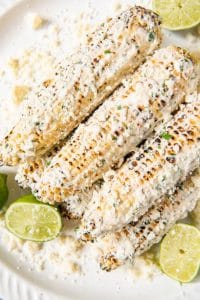 An image of a stack of ears of corn on the cobb made mexican-style with mayo, sour cream, lime, cilantro, chili powder, garlic, and cotija cheese, surrounded by sliced limes and extra crumbled cheese.