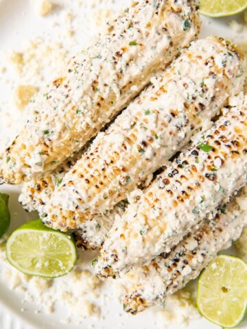 An image of a stack of ears of corn on the cobb made mexican-style with mayo, sour cream, lime, cilantro, chili powder, garlic, and cotija cheese, surrounded by sliced limes and extra crumbled cheese.
