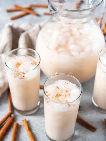 A pitcher of ice cold horchata next to cups of horchata and sticks of cinnamon