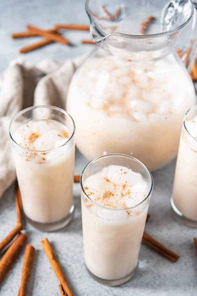 A pitcher of ice cold horchata next to cups of horchata and sticks of cinnamon