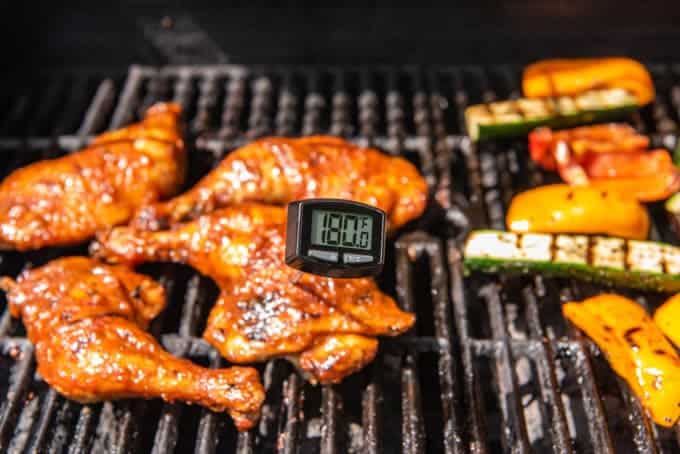 An image of an instant read meat thermometer at 180 degrees F for barbecue chicken legs and thighs on the grill, with grilled vegetables on the side.