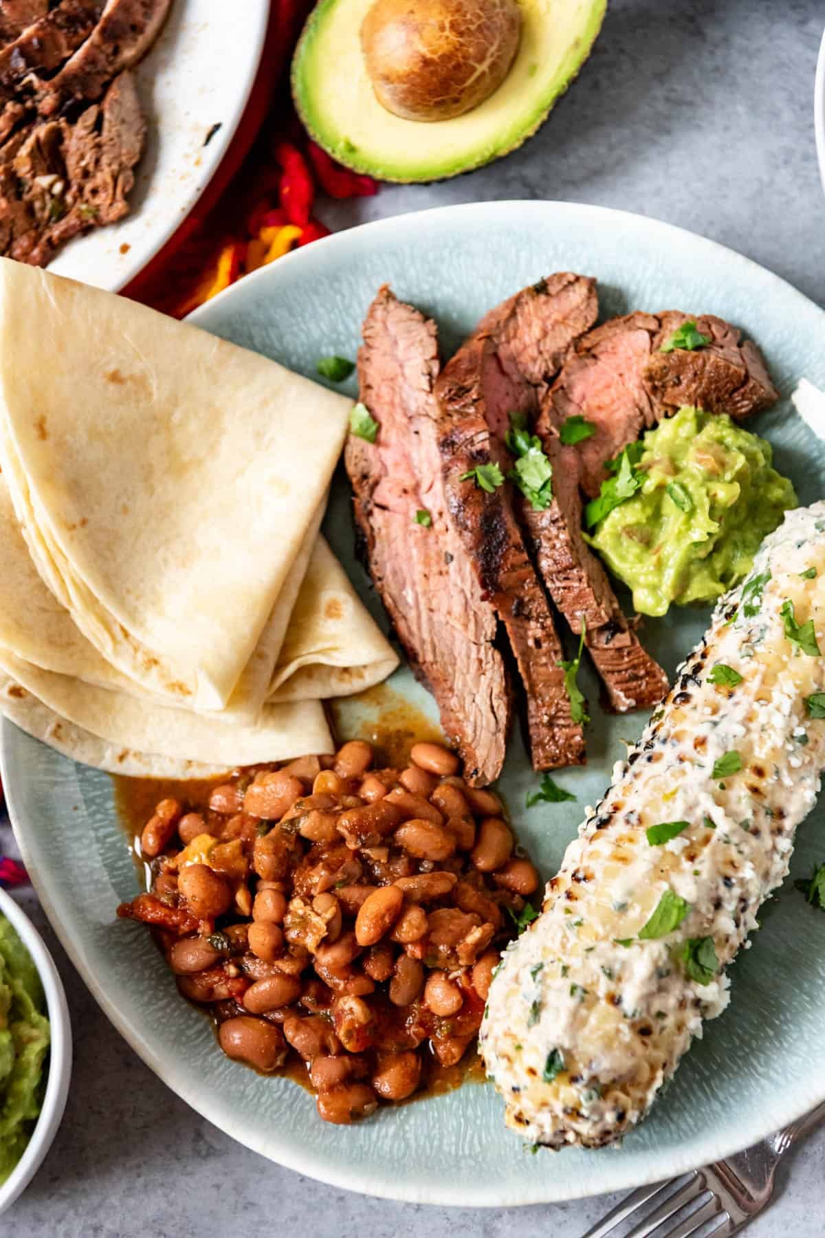 An image with a full mexican dinner for cinco de mayo with charro beans, flour tortillas, grilled Mexican street corn, and carne asada sliced into thin strips.