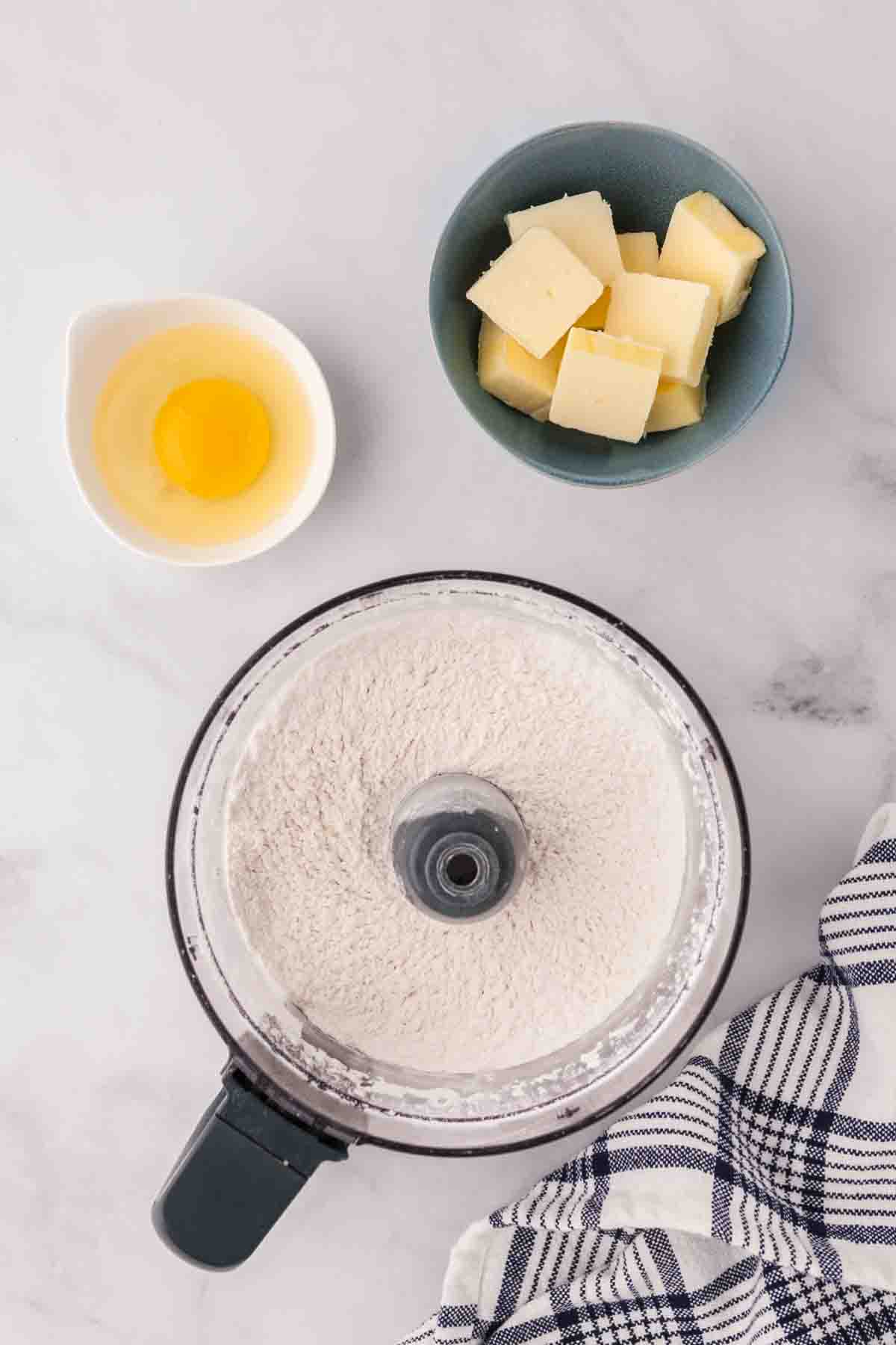 Adding flour to a food processor with an egg and butter nearby.