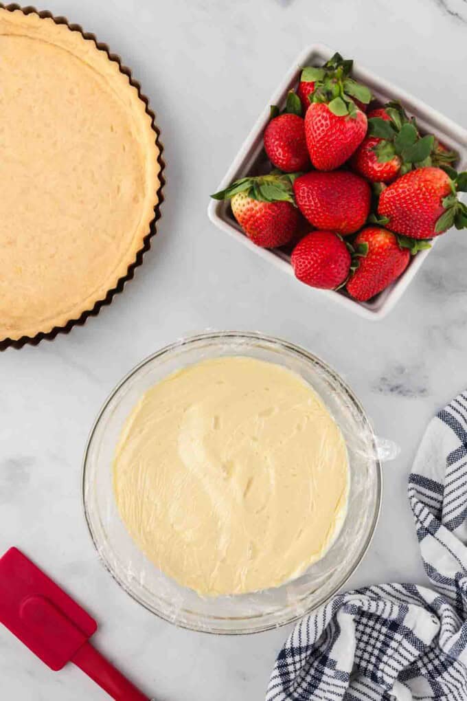 Chilled pastry cream in a bowl next to a tart crust and a container of strawberries.