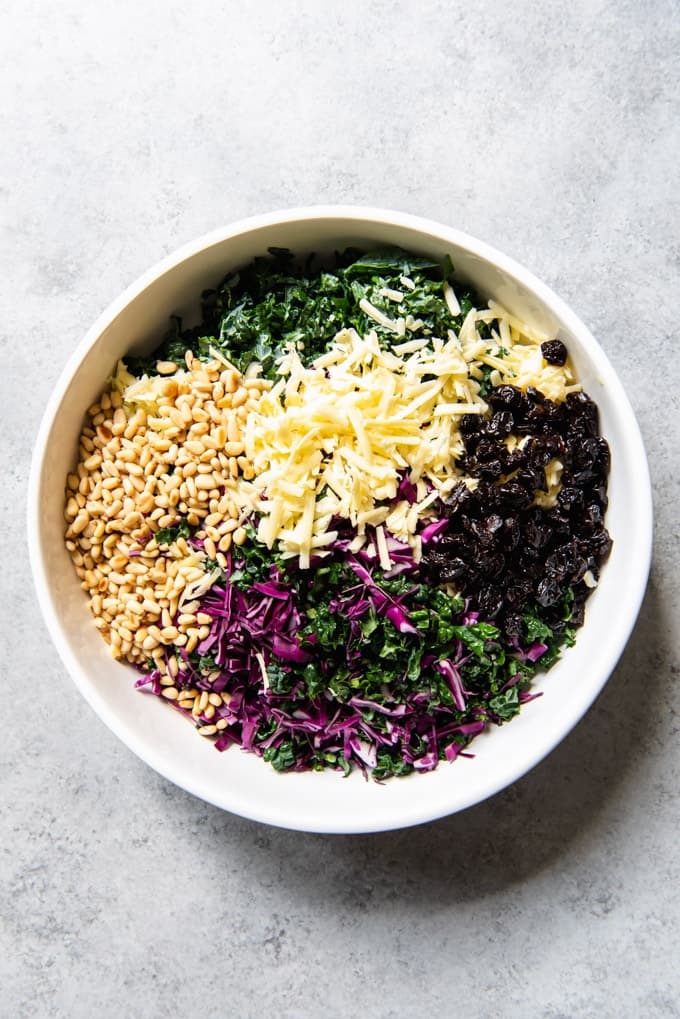 An image of a large white serving bowl with chopped kale, red cabbage, white cheddar cheese, pine nuts, and dried cherries for a chopped kale salad.