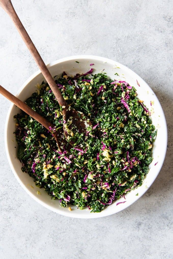 A large white bowl of chopped kale salad with two serving spoons in it.