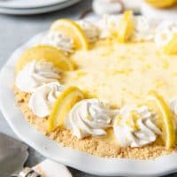 a no bake sour cream lemon pie in a white pie plate garnsihed with whipped cream and lemon slices