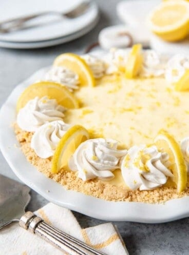 a no bake sour cream lemon pie in a white pie plate garnsihed with whipped cream and lemon slices
