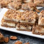 a white plate filled with stacked pecan pie cheesecake bars with scattered whole pecans below