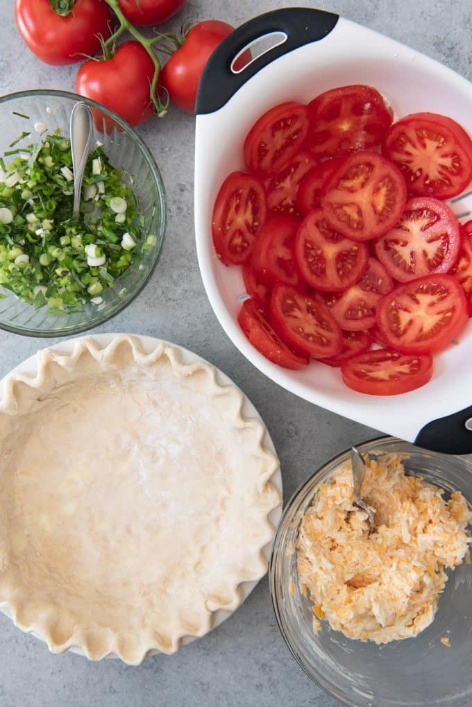 An image of the ingredients for a tomato pie - pie crust, tomatoes, cheese, mayo, green onions, and basil.