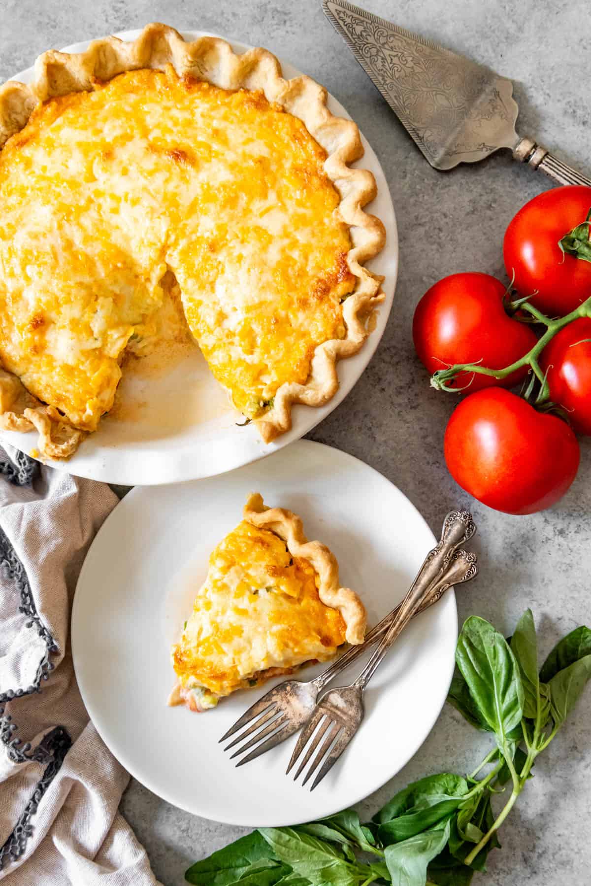 An image of a cheese and tomato pie with one slice removed and set on a white plate next to summer-ripe tomatoes and fresh basil.