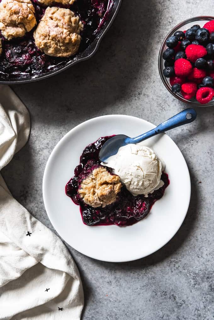 An image of a plate of mixed berry cobbler with a biscuit topping served with a scoop of vanilla ice cream on the side.