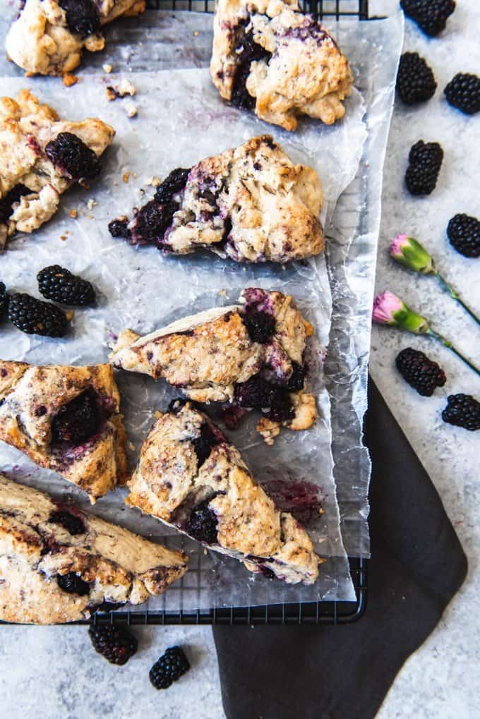 An image of fruit scones made with blackberries, set on top of waxed paper and a cooling rack with blackberries sprinkled around them.
