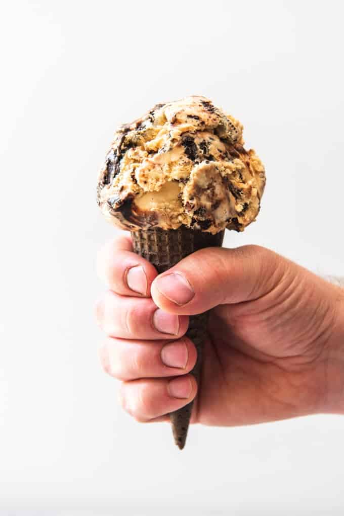 An image off a hand holding a chocolate sugar cone with a scoop of caramel oreo fudge ripple ice cream.
