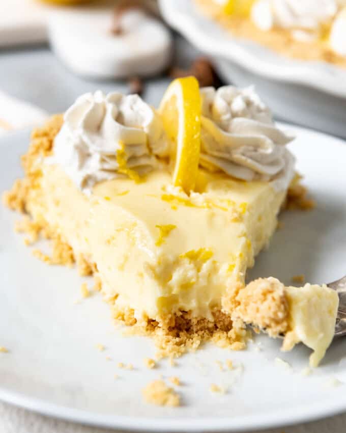 An image of a slice of lemony no-bake sour cream lemon pie with swirls of whipped cream and a slice of lemon on top, with a bite taken out of it.