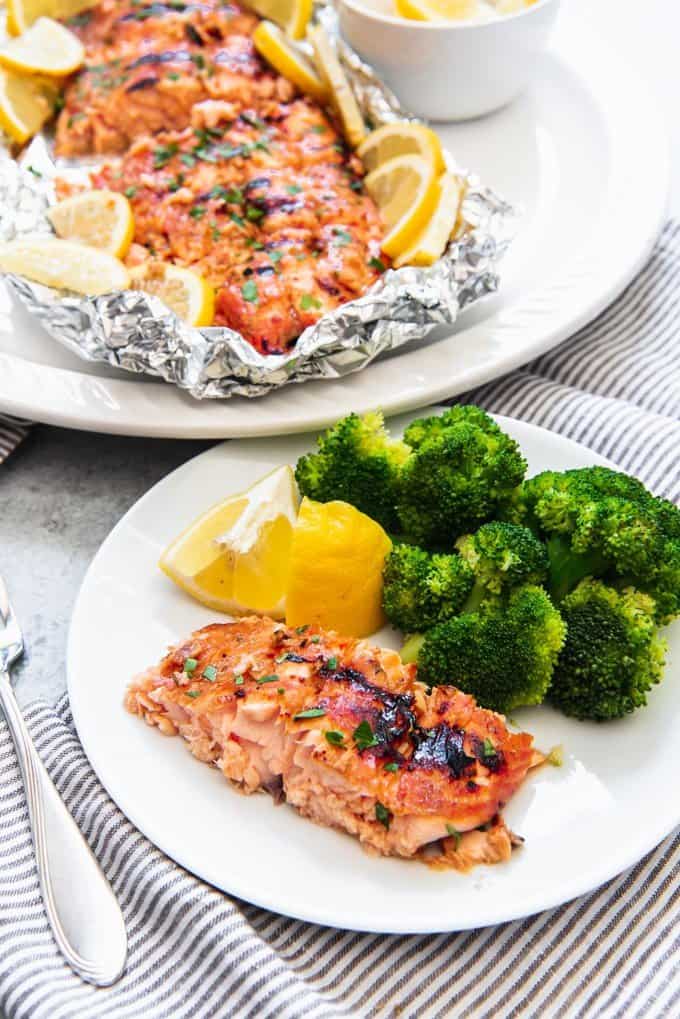 An image of an individual portion of grilled salmon in foil on a white plate with steamed broccoli and lemon wedges.