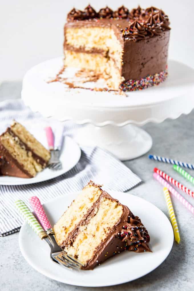 two slices of yellow cake with chocolate frosting below a cakestand of chocolate frosted yellow cake