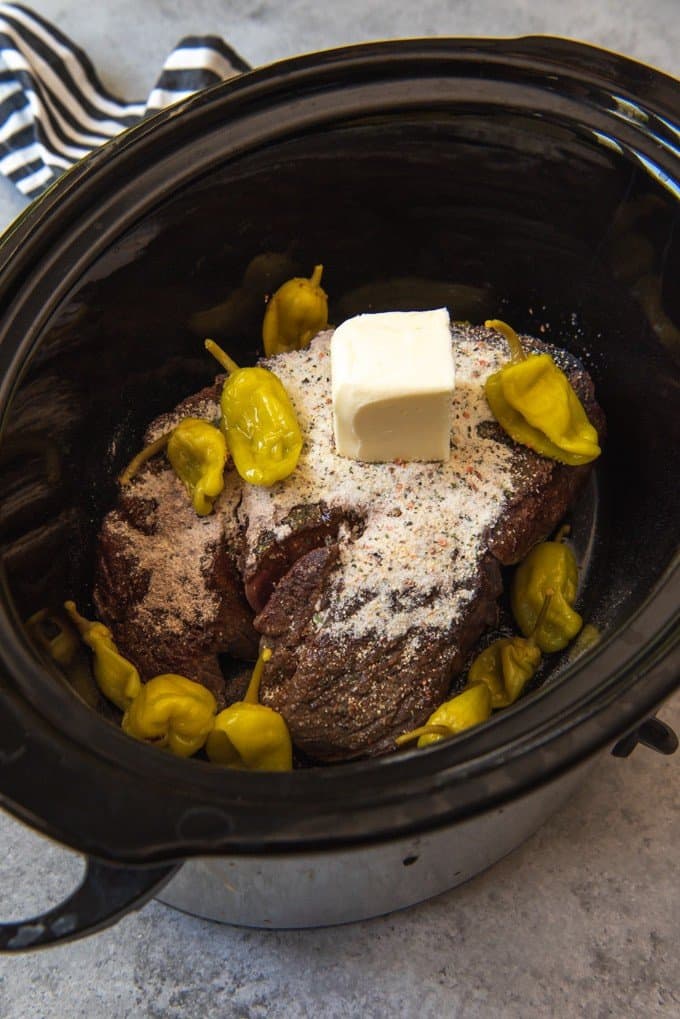 An image of a browned roast in a crock pot with pepperoncini peppers, italian seasoning, au jus gravy mix, and butter for Mississippi pot roast sandwiches.