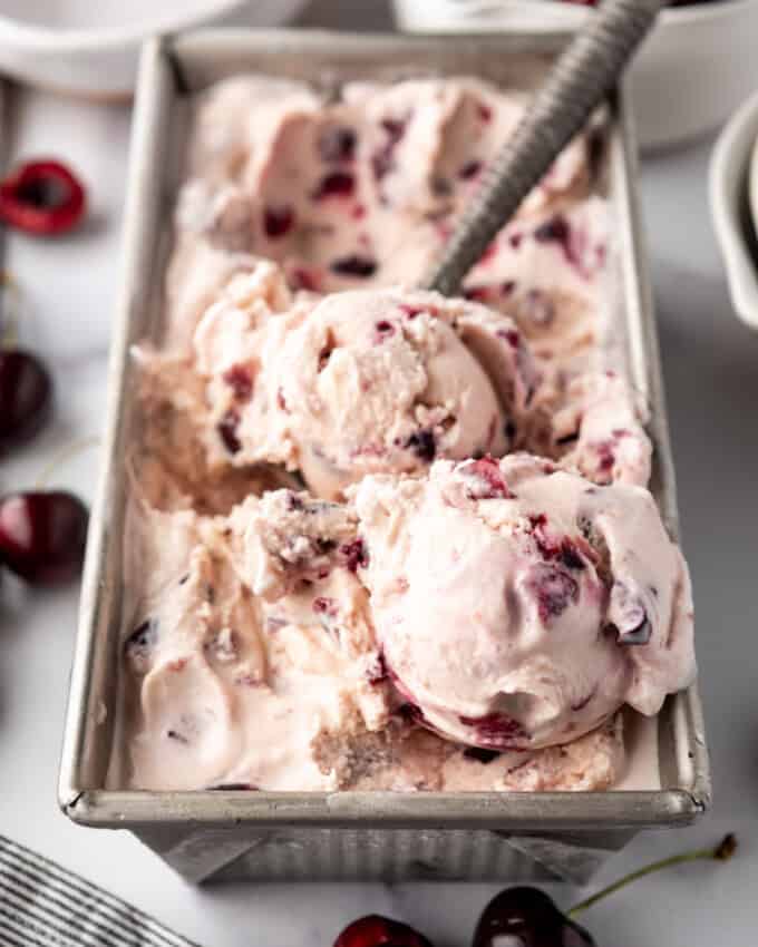 An image of a container of homemade cherry vanilla ice cream with a bowl of cherries and an ice cream scoop nearby.