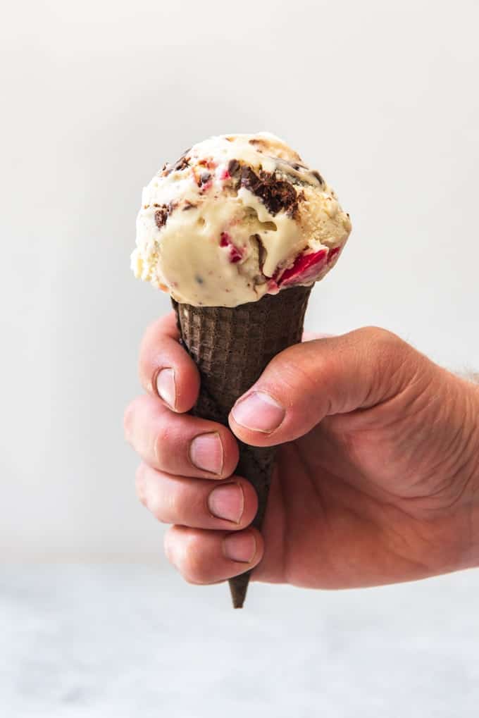 An image of a hand holding an ice cream cone with a scoop of white chocolate ice cream with a raspberry swirl and chocolate truffle pieces.