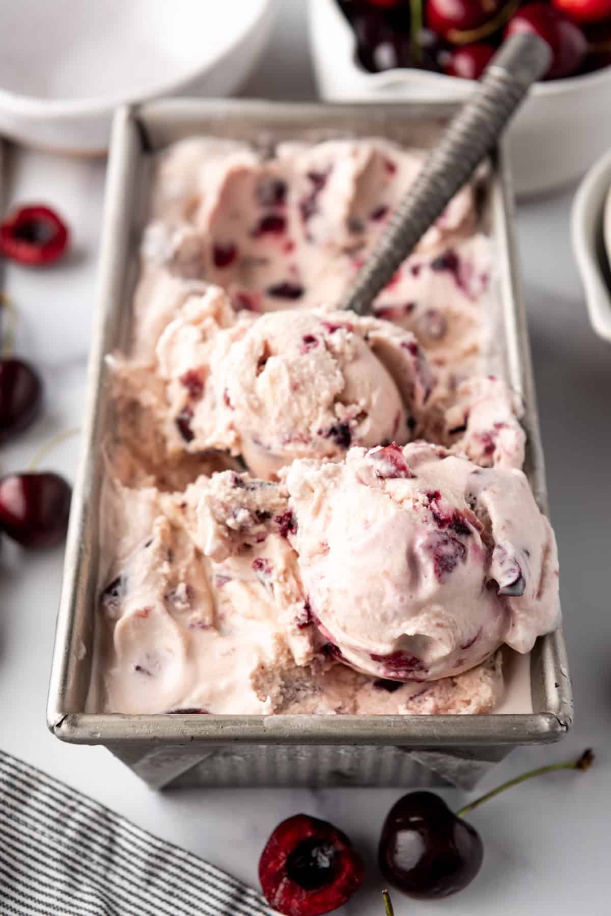 An image of a container of homemade cherry vanilla ice cream with a bowl of cherries and an ice cream scoop nearby.