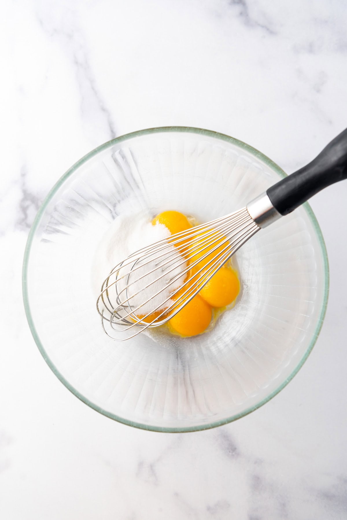 A whisk in a glass bowl with egg yolks and sugar.