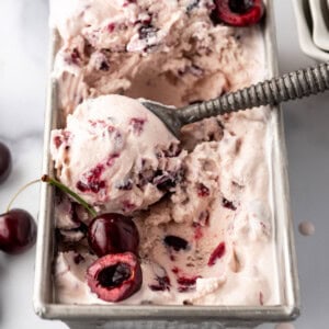 An image of a container of homemade cherry vanilla ice cream with a bowl of cherries and an ice cream scoop nearby.