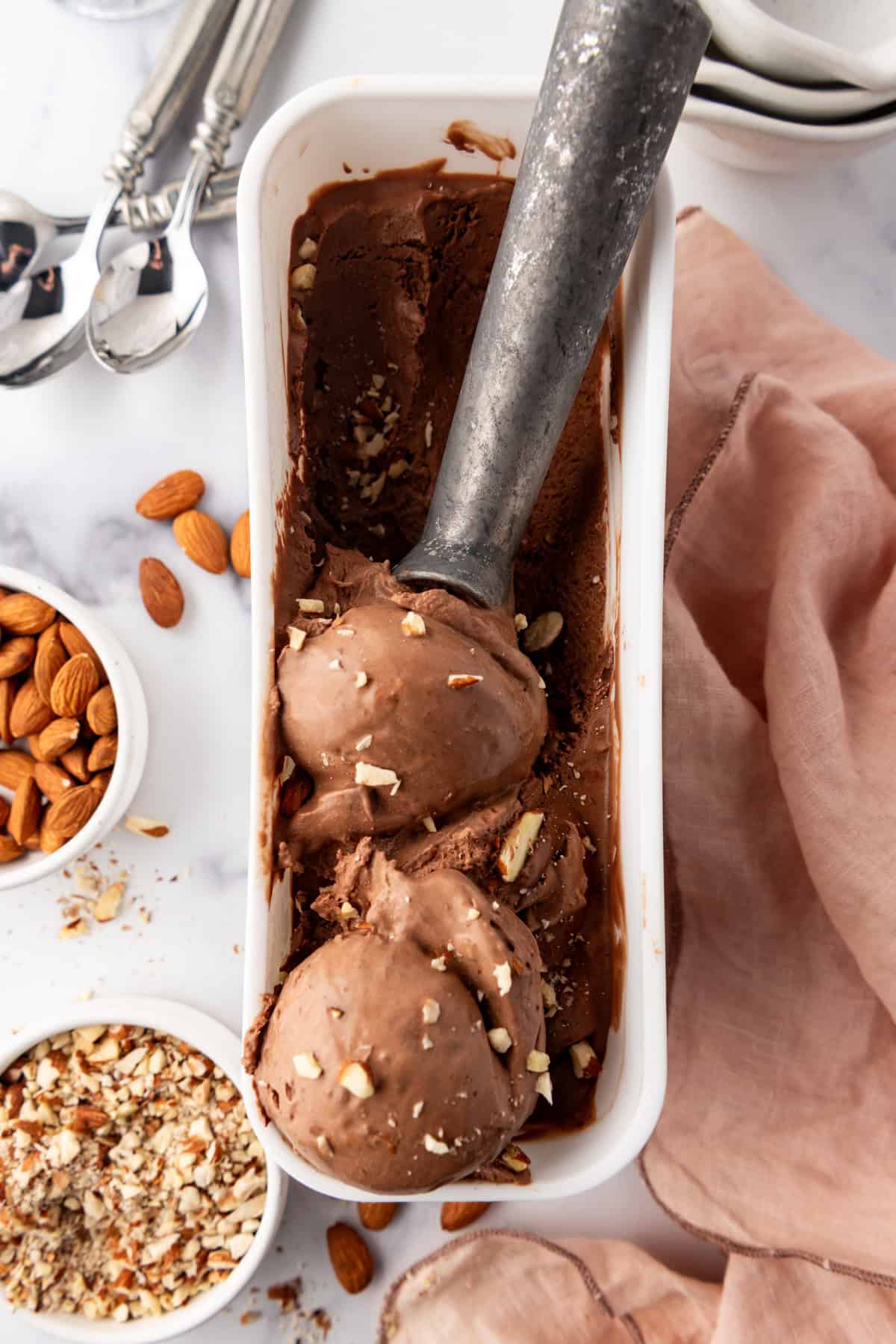 An overhead image of scoops of burnt almond fudge ice cream in a white container next to chopped almonds.