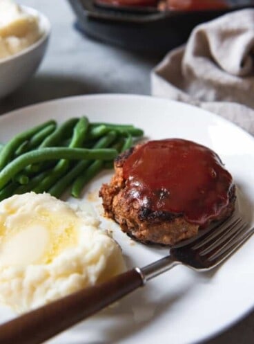 meatloaf pattie on a white plate with green beans mashed potatoes and a fork