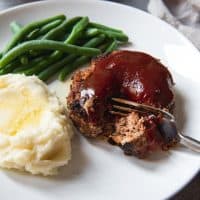 An image of a meatloaf patty with a bite taken out of it on a white plate with green beans and a pile of mashed potatoes dripping with melted butter.
