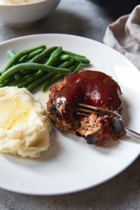 An image of a meatloaf patty with a bite taken out of it on a white plate with green beans and a pile of mashed potatoes dripping with melted butter.