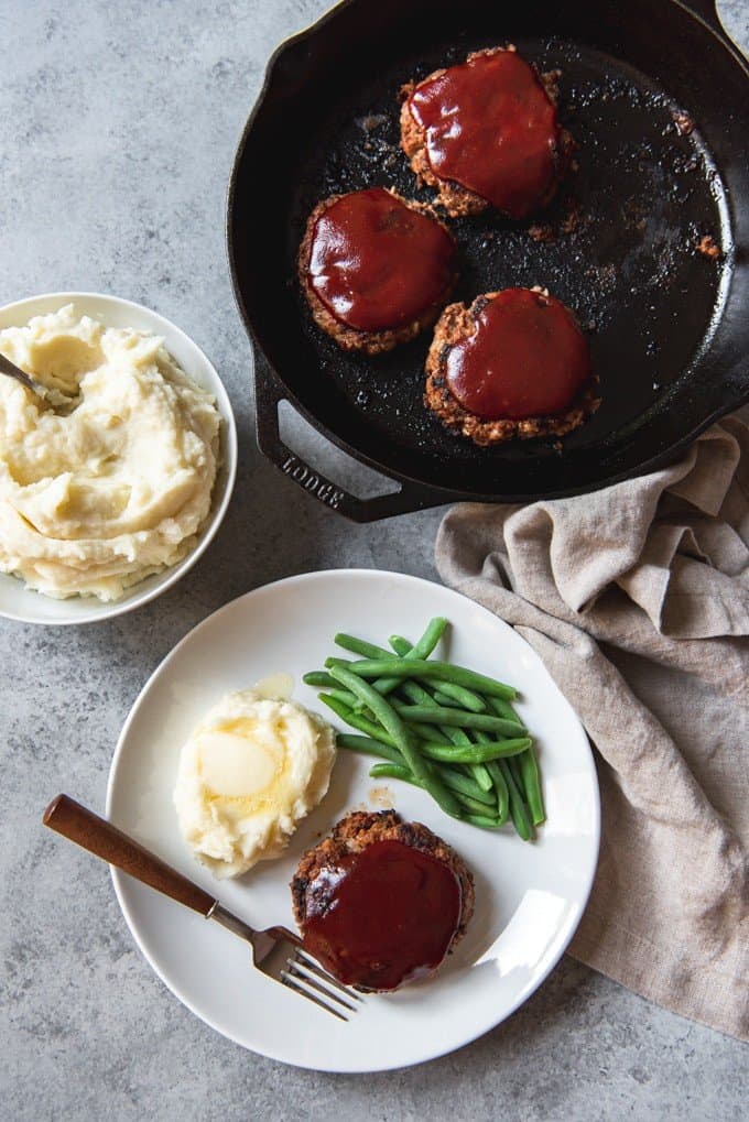 An image off an individually-sized meatloaf patty on a plate with mashed potatoes, green beans, and a skillet beside it with more meatloaf patties.
