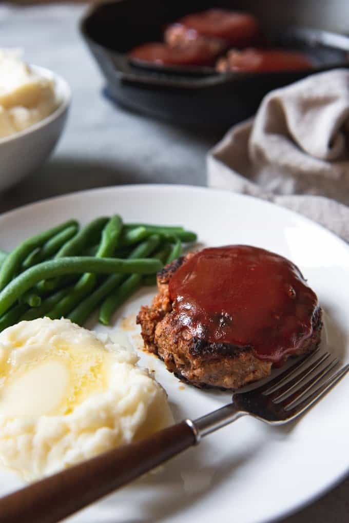 meatloaf pattie on a white plate with green beans mashed potatoes and a fork