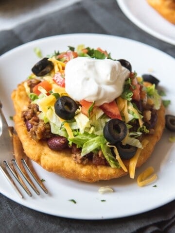 a frybread taco on a white plate next to a fork