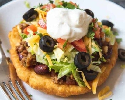 a frybread taco on a white plate next to a fork