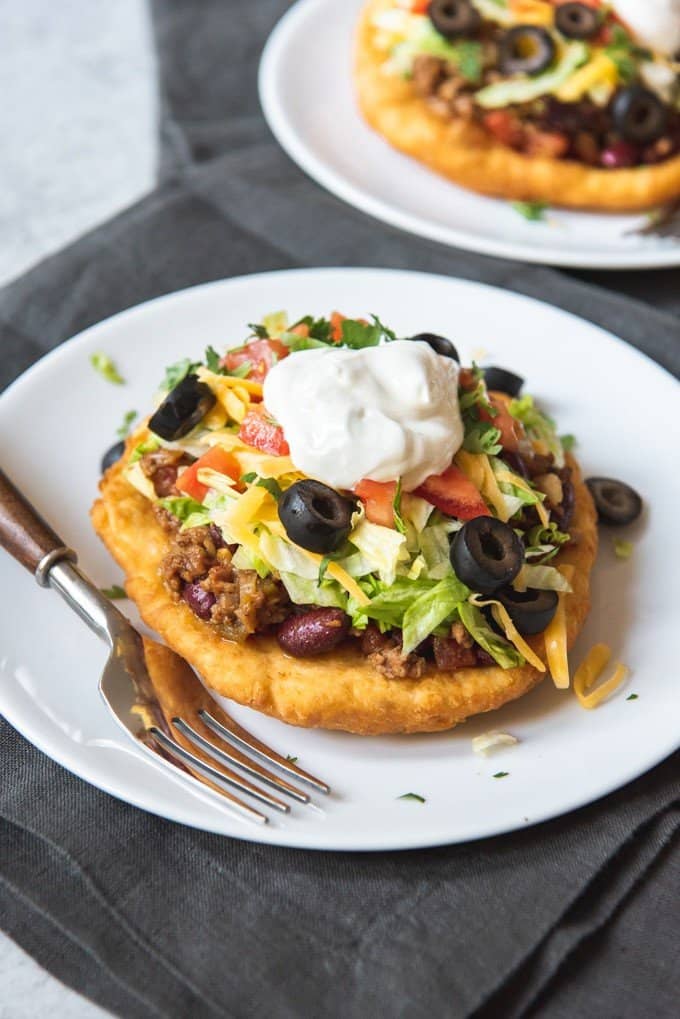 An image of Indian Tacos made with Indian fry bread, then topped with taco meat, beans, lettuce, tomatoes, cheese, olives, and sour cream.