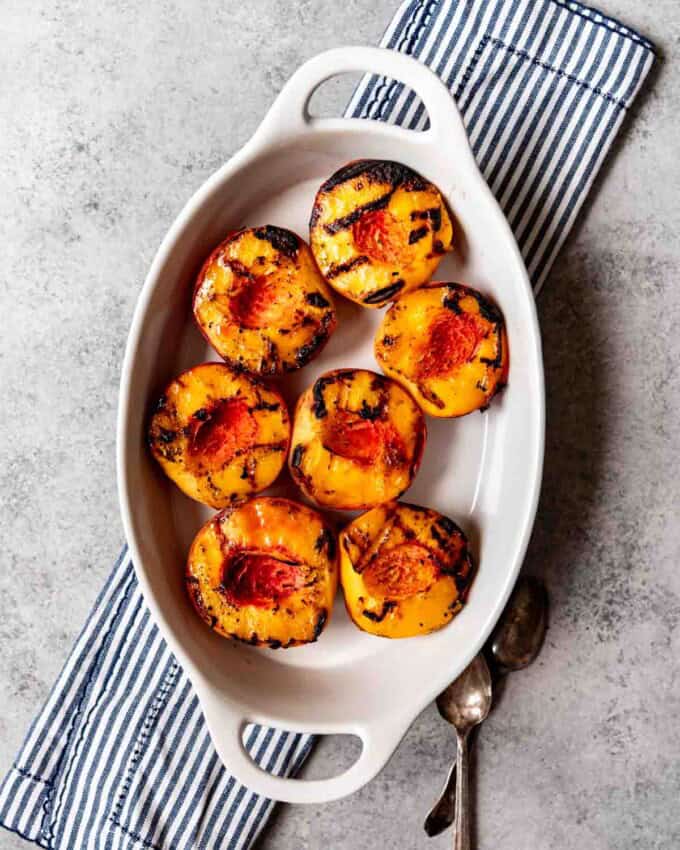 An aerial view of a white baking dish with 7 grilled peach halves inside.