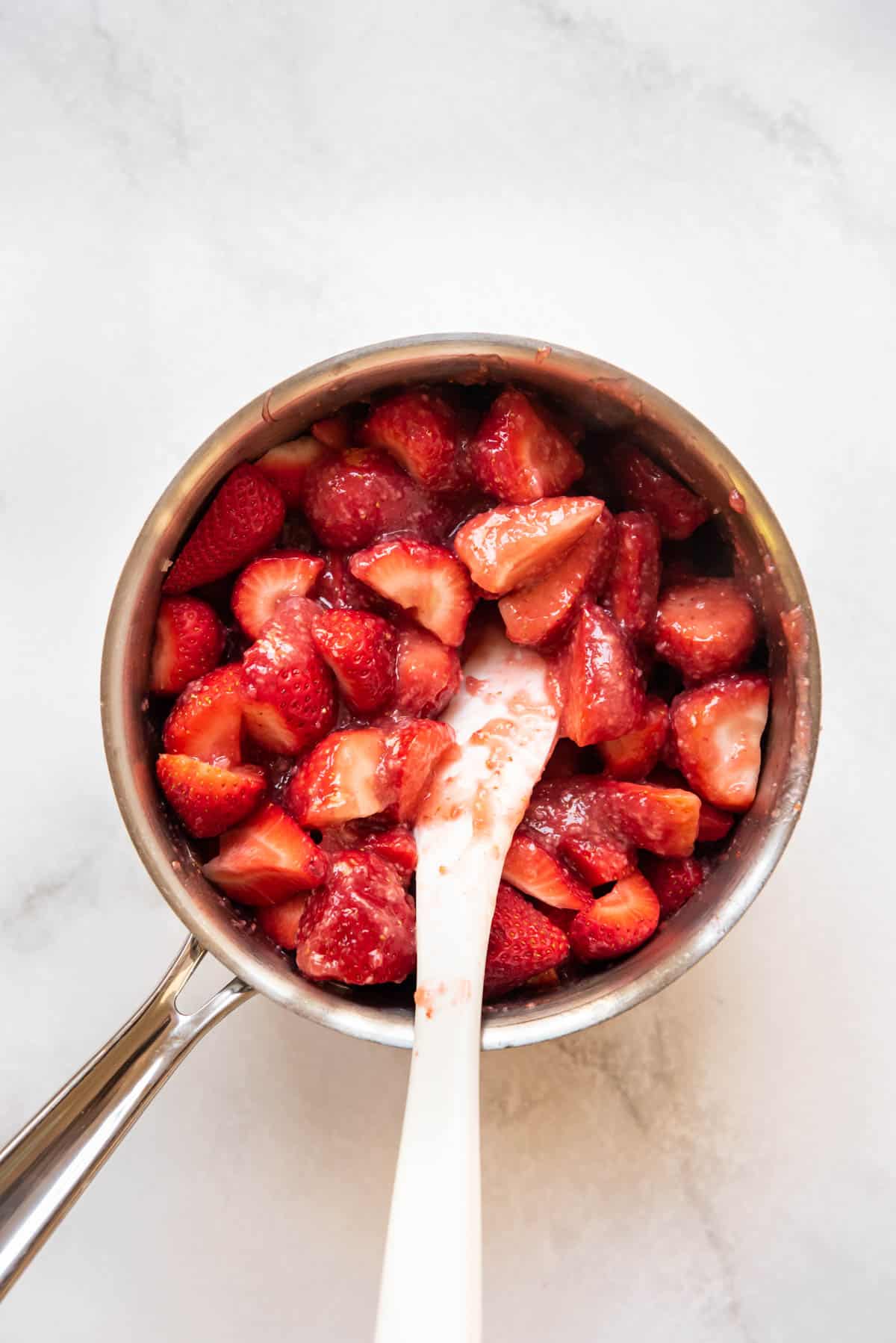 Stirring strawberries into strawberry pie glaze made from scratch in a saucepan.