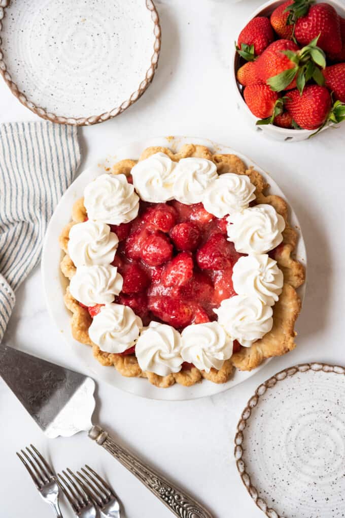 An overhead image of a fresh strawberry pie with piped swirls of whipped cream decorating the top next to a bowl of strawberries.