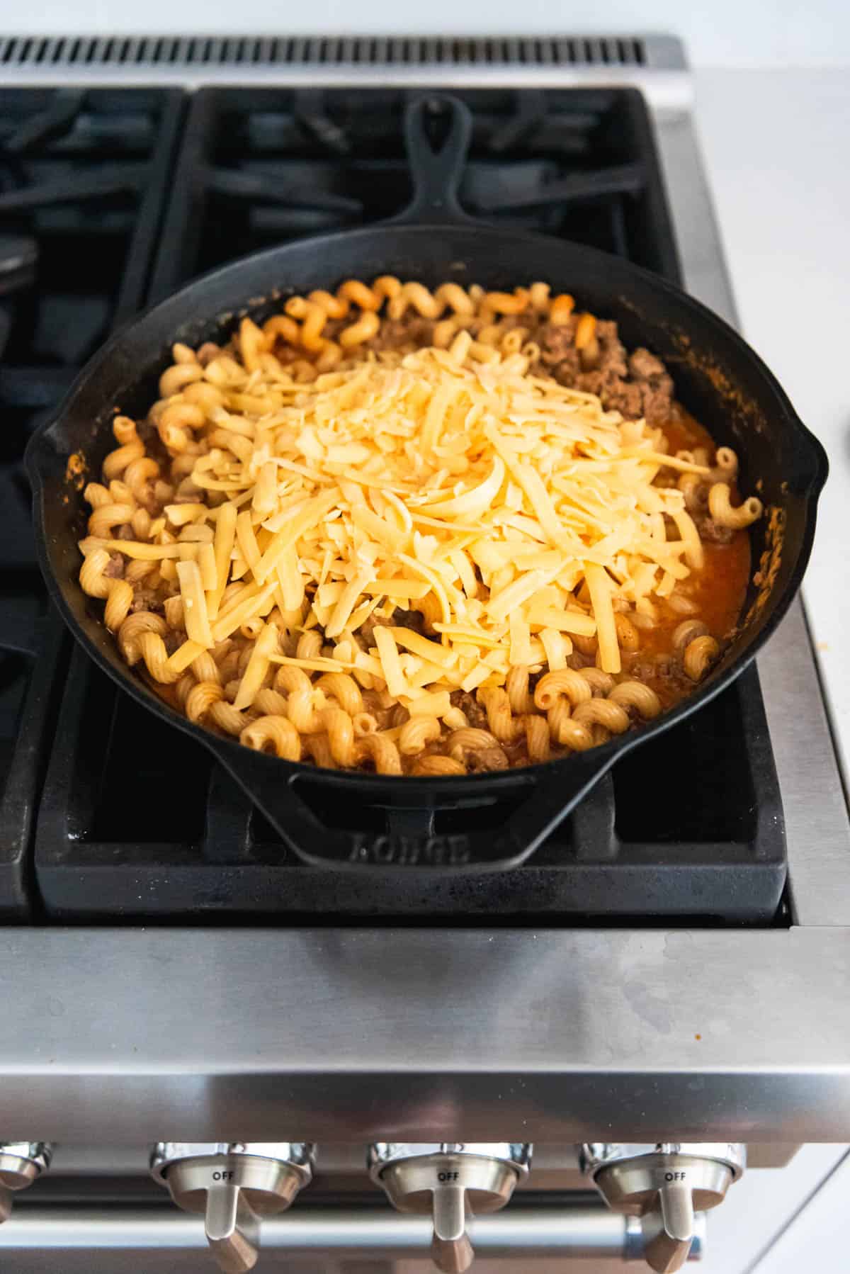 An image of shredded cheese being added to cooked pasta and ground beef in a cast iron skillet.