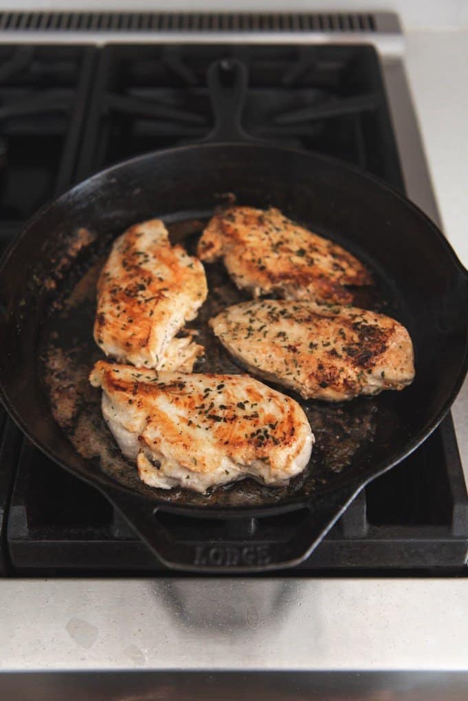 An image of four chicken breasts being seared in a cast iron skillet on the stovetop.