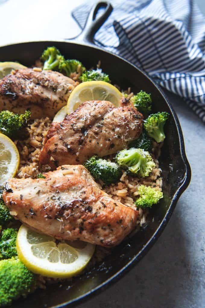 An image of golden brown seared chicken breasts in a skillet with broccoli, rice, and lemon wedges.