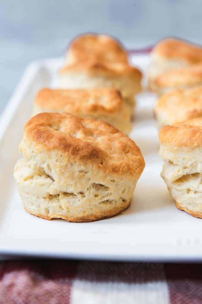 An image of flaky, fluffy, tall buttermilk biscuits with golden tops on a white baking sheet.