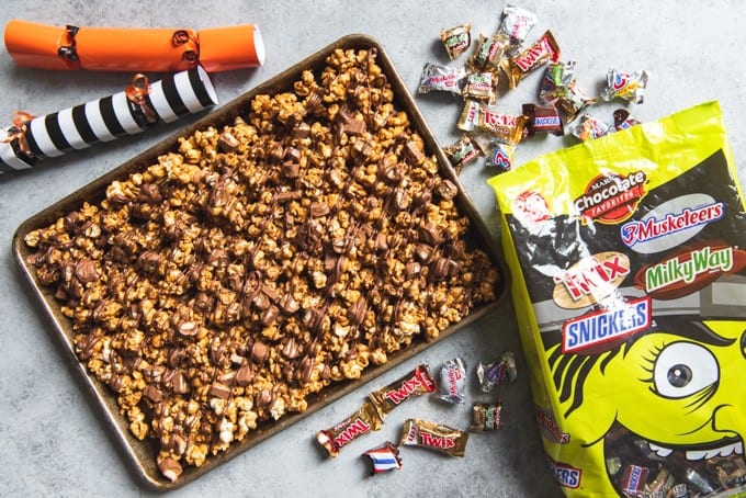 An image of a tray of homemade caramel popcorn made with Twix candy bars, surrounded by mini candies and Halloween poppers.
