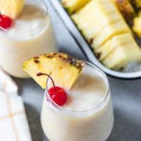 virgin pina colads in small glasses with pineapple wedges and cherries for garnish and a tray of sliced pineapple in back