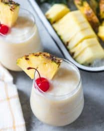virgin pina colads in small glasses with pineapple wedges and cherries for garnish and a tray of sliced pineapple in back