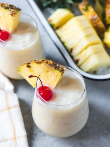 virgin pina colads in small glasses with pineapple wedges and cherries for garnish and a tray of sliced pineapple in back