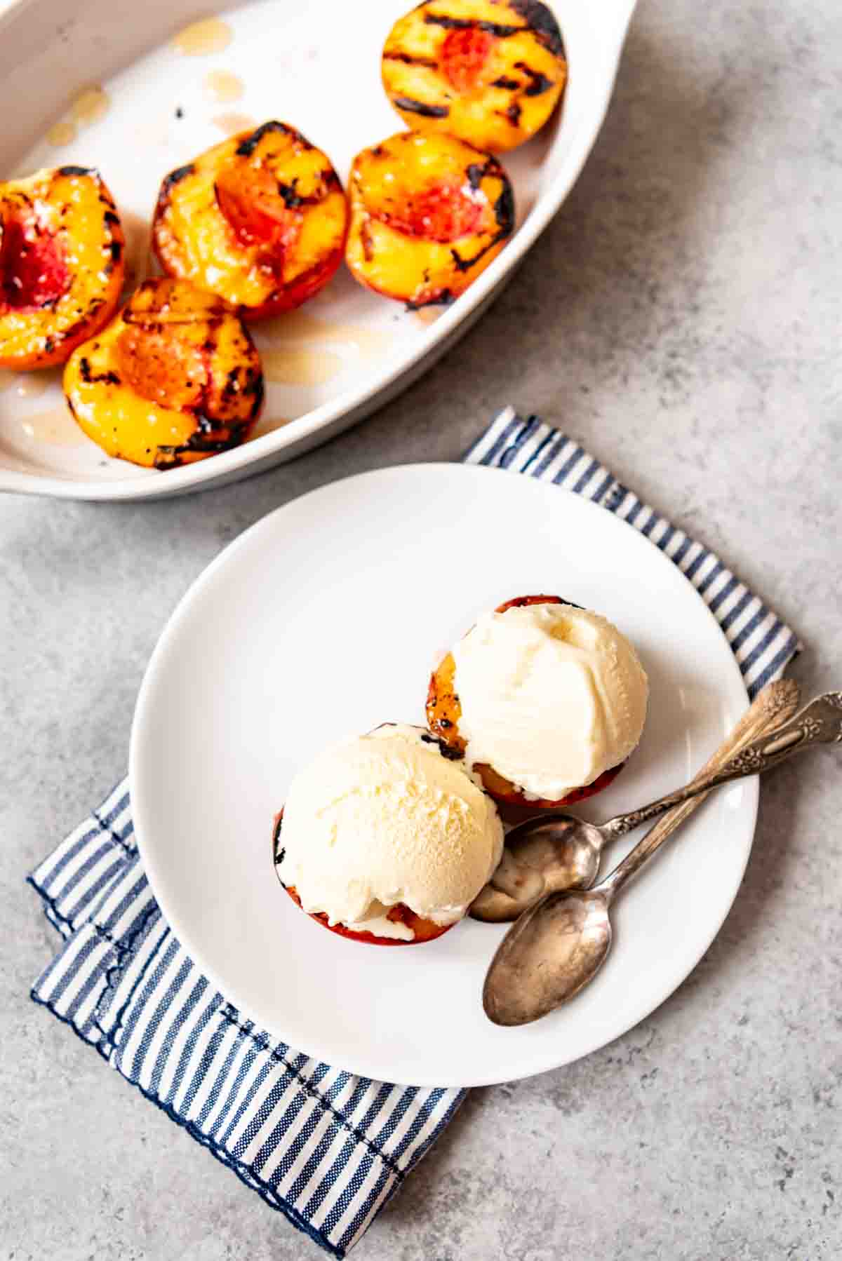 An overhead image of grilled peaches with ice cream on a white plate with spoons.