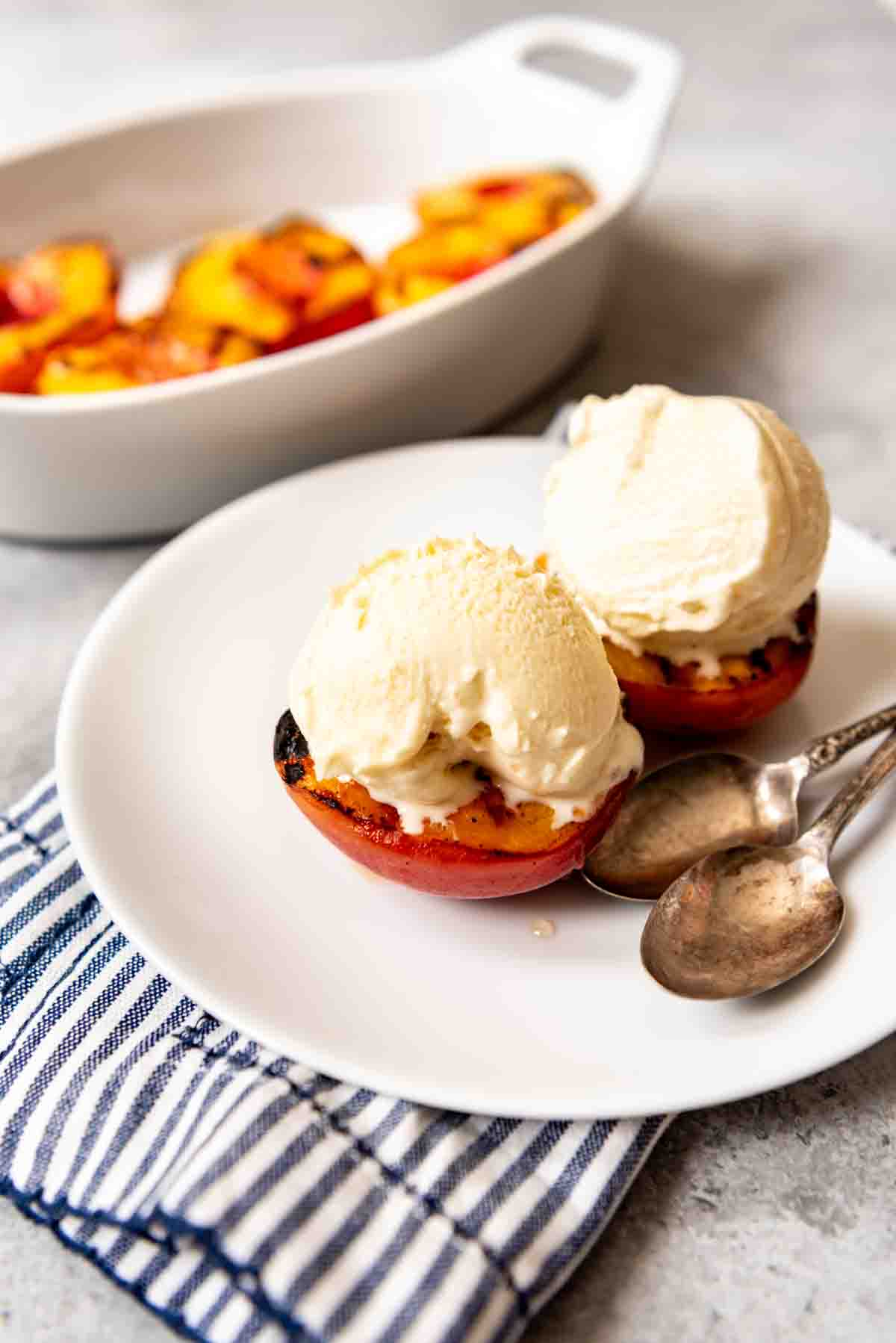 Two grilled peaches with ice cream on top on a white plate with two spoons.