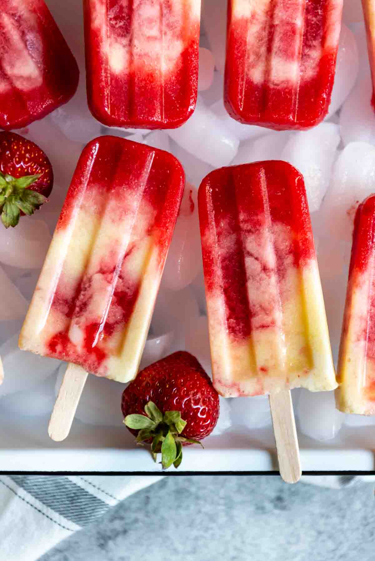 An image of two strawberry pineapple coconut swirl popsicles side by side on a bed of ice cubes.